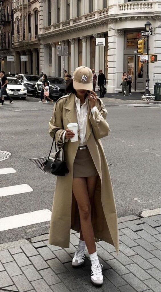 Fashionable woman in a beige trench coat, New York cap, and sneakers, walking in city with coffee. Urban chic style.