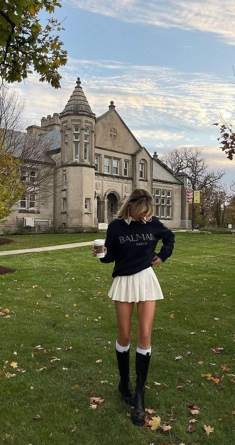 Person in stylish outfit holding coffee on a grassy lawn in front of a historic mansion under a blue sky.