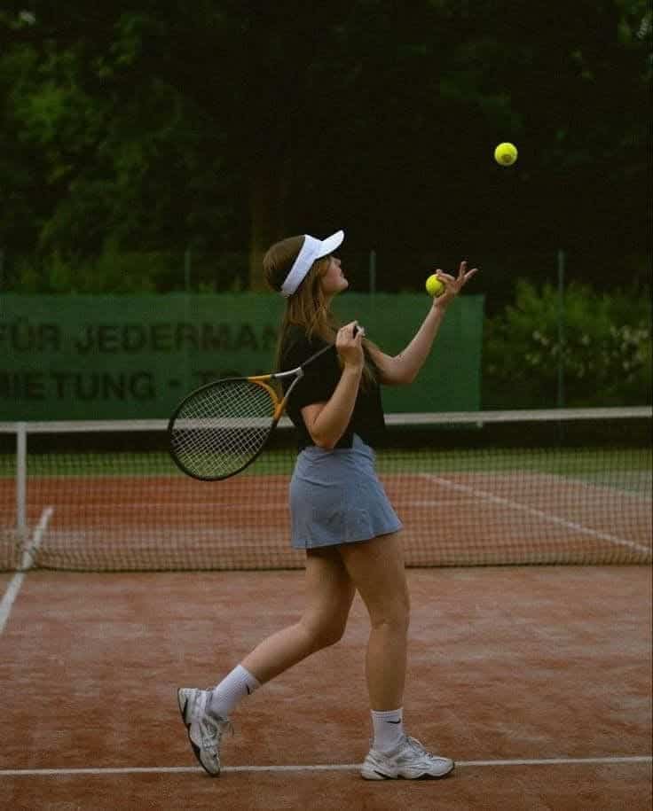 Tennis player juggling balls on clay court, holding racket, wearing visor and sports attire, in action shot.