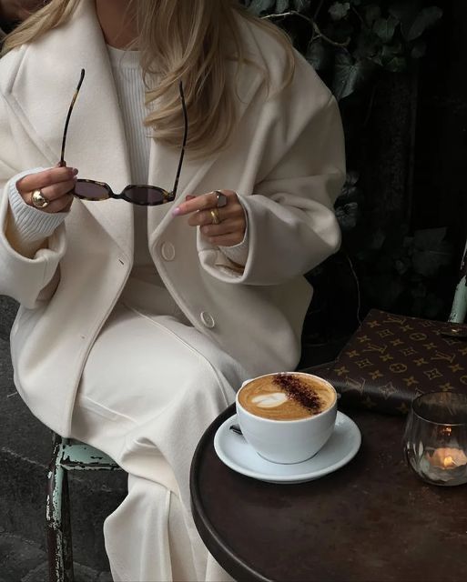 Stylish woman in cream coat with sunglasses, enjoying cappuccino at outdoor café table.
