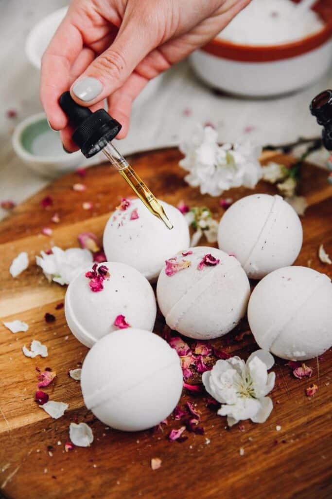 Hand adding essential oil to bath bombs with rose petals on wooden board for relaxation and skincare.