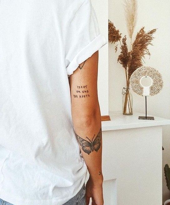Person with butterfly tattoo on arm, wearing a white shirt in minimalistic room with dried plants and modern decor.