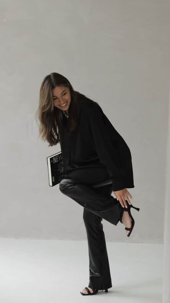 Woman in stylish black outfit adjusting shoe, smiling indoors. Fashionable look with leather pants and sandals.