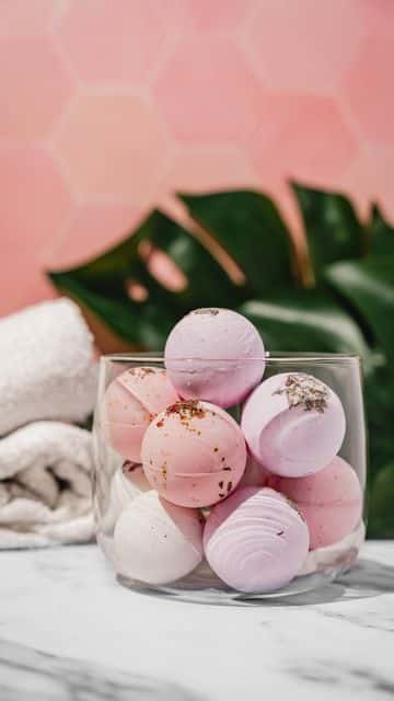 Glass bowl with pink bath bombs, against a pink hexagonal backdrop, white towels, and lush green leaves.