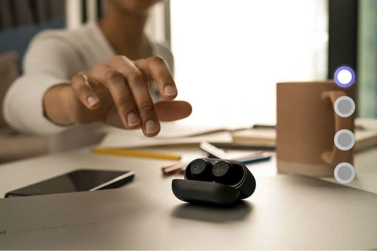 Hand reaching for wireless earbuds on a desk with a smartphone, pencils, and coffee mug in background.