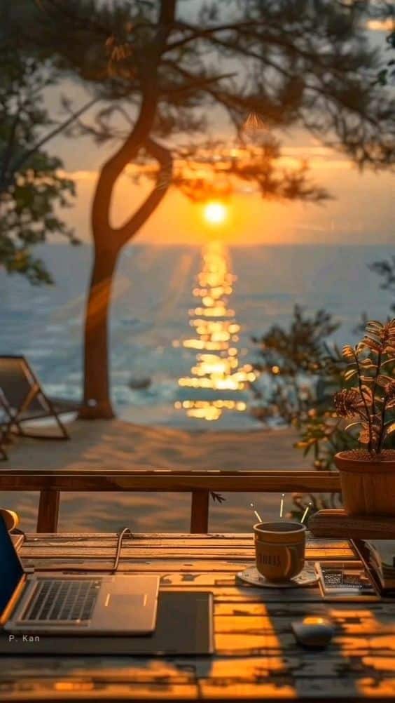 Laptop and coffee on a wooden desk with a scenic ocean sunset view, creating a peaceful work setting.