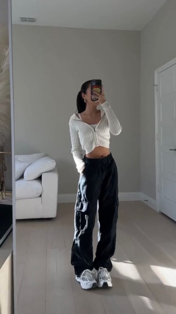 Person taking a mirror selfie in casual white top and black cargo pants, standing in a stylishly minimal room.