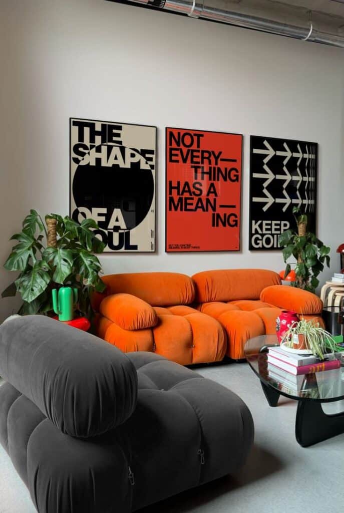 Modern living room with bold poster art, orange and gray tufted sofas, plants, and a glass coffee table.