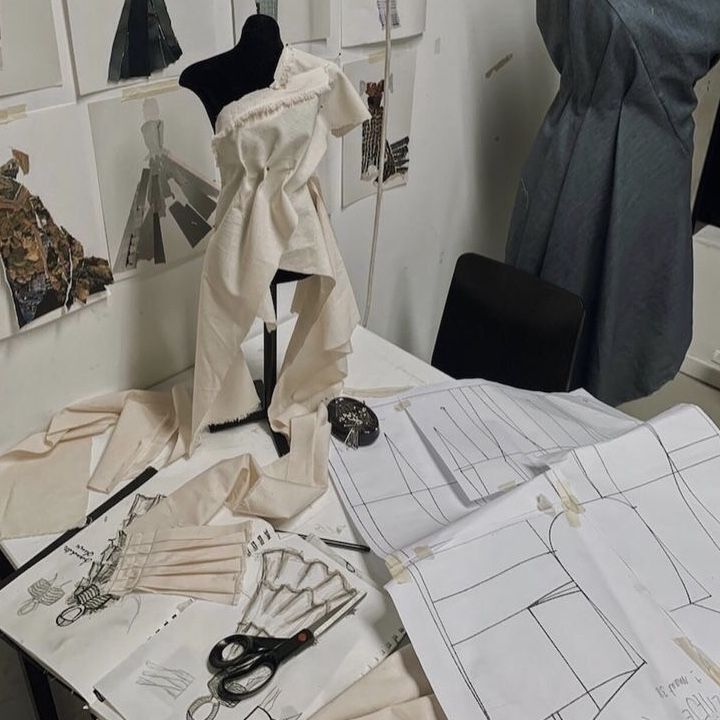 Fashion design workspace with fabric draped on mannequin, sewing patterns, and sketches on a cluttered table.