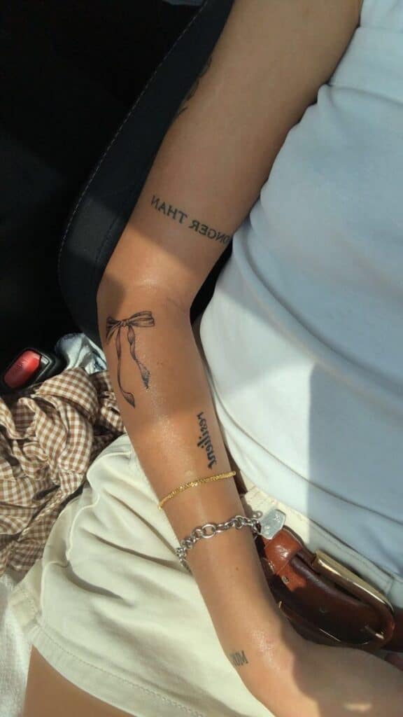 Close-up of a person’s arm with tattoos and wearing a white top, showcasing a bow and text tattoo.