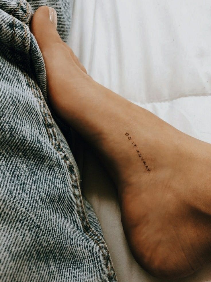 Close-up of a minimalist ankle tattoo reading Do it anyway, next to denim fabric, emphasizing personal expression.