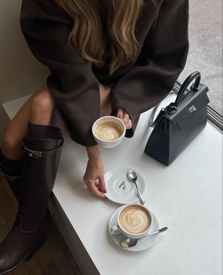 Woman in a cozy setting enjoying coffee by the window, wearing boots, with a handbag on the table.