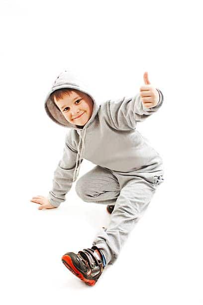 Child in gray hoodie gives thumbs up while kneeling, smiling playfully.