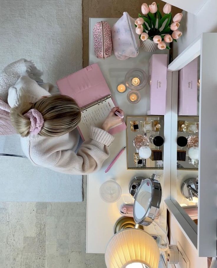 Woman at pink-themed desk writing in notebook with candles and tulips nearby. Cozy workspace ambiance.
