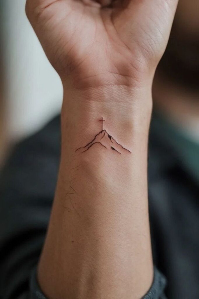 Minimalist mountain tattoo on a wrist with a small cross on the peak, symbolizing faith and adventure.