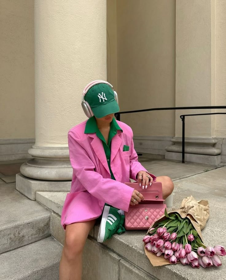 Stylish woman in pink blazer and sneakers, wearing headphones, sits with a handbag and tulips outside a classic building.