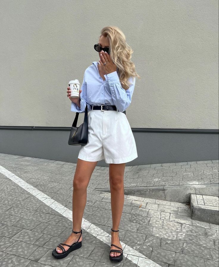 Fashionable woman with coffee, wearing a blue shirt and white shorts, stands stylishly on a paved surface.