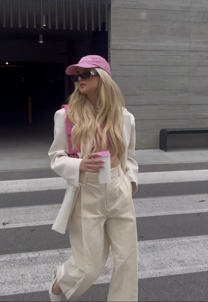 Fashionable woman in pink cap and sunglasses crossing street with coffee, wearing cream outfit and white blazer. Summer Outfit