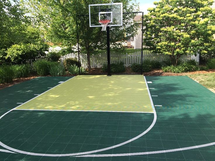 Outdoor home basketball court with green and yellow surface, surrounded by lush trees and a white picket fence.