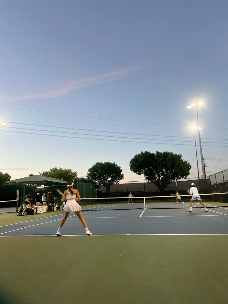 Tennis players compete in a doubles match at dusk on a lit court, with trees and a clear sky in the background.
