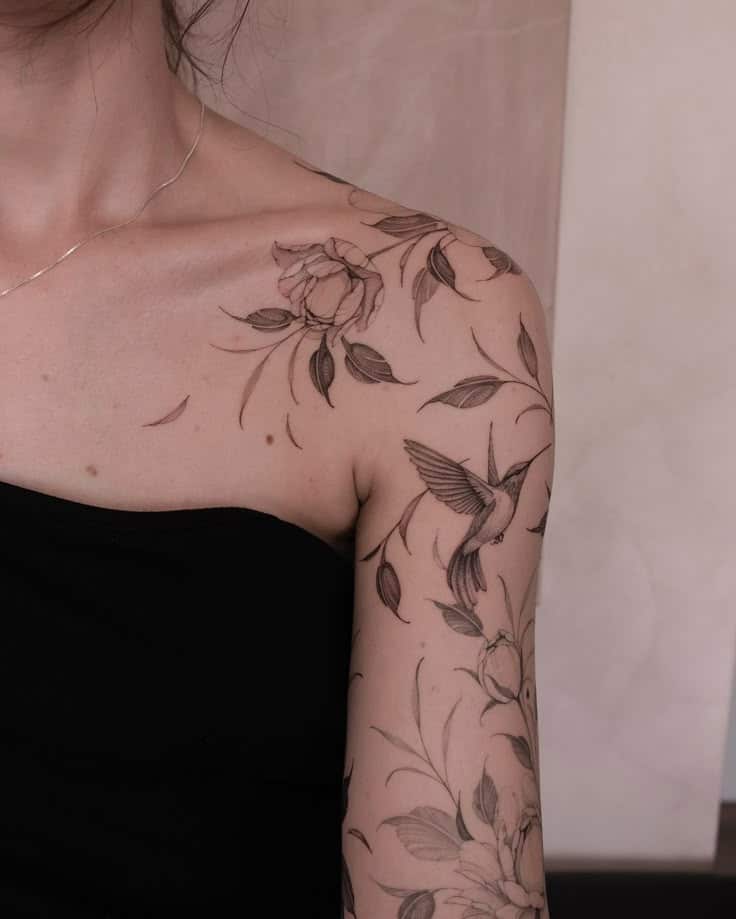 Delicate floral and hummingbird tattoo on a woman's shoulder and arm, wearing a black top.