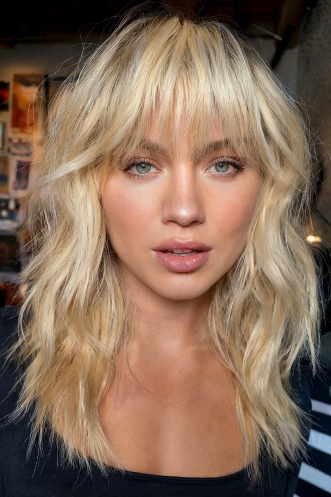 Blonde woman with wavy hair and bangs, wearing a black top, gazing forward with a layered hairstyle in a casual setting. Hair Styles
