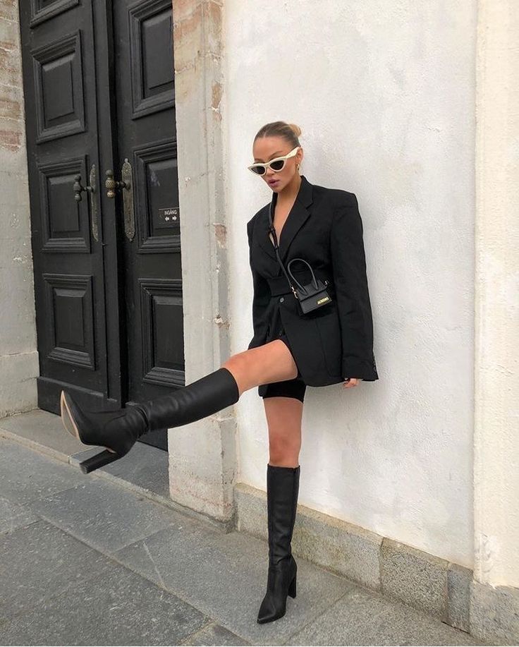 Fashionable woman in black outfit and boots posing confidently against a wall.