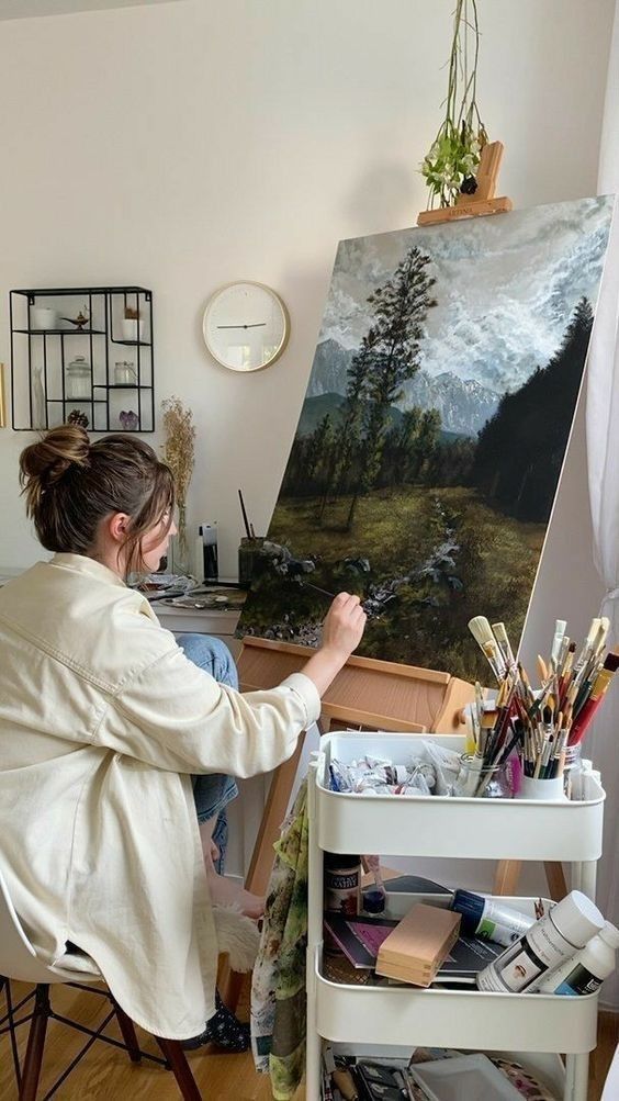 Artist painting a serene mountain landscape on canvas in a cozy studio filled with art supplies.