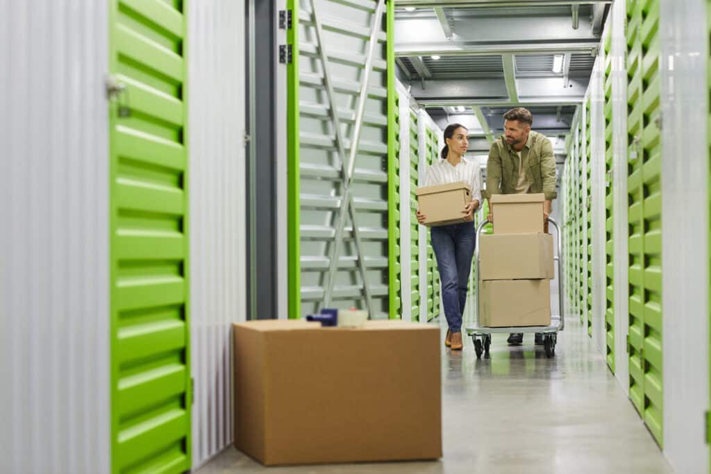 Two people carrying boxes in a bright storage facility with green doors.