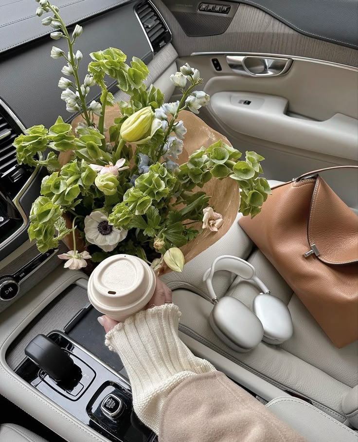 Coffee cup, fresh flowers, headphones, and a leather bag on a car seat for a cozy travel vibe.
