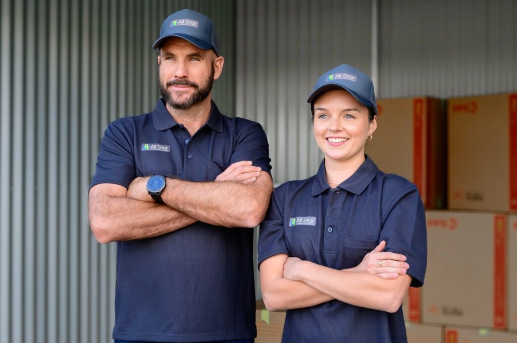 Two delivery employees in branded uniforms posing confidently in front of stacked boxes.