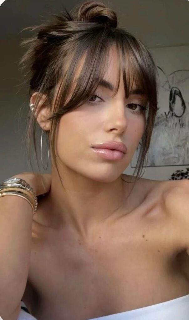 Woman with updo hairstyle and bangs, wearing hoop earrings and bracelets, poses confidently in a casual indoor setting.