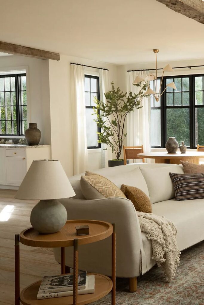 Cozy living room with a beige sofa, wooden side table, large windows, and indoor plants for a modern, airy vibe.