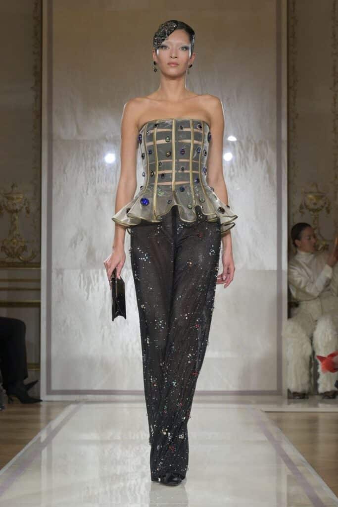 Model showcasing an embellished corset top with sheer pants on the runway, fashion show setting.