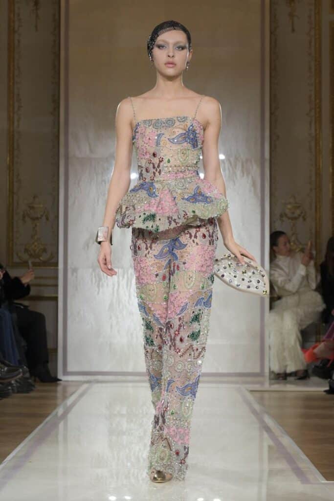 Fashion model in colorful, embellished outfit with peplum detail walking runway at elegant indoor fashion show.