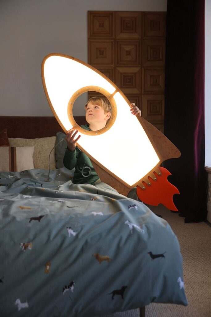 Child holding a glowing rocket-shaped lamp while seated on a bed in a cozy, decorated room.