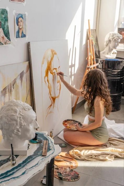 Artist painting portrait on canvas in sunlit studio, surrounded by sculptures and art supplies.