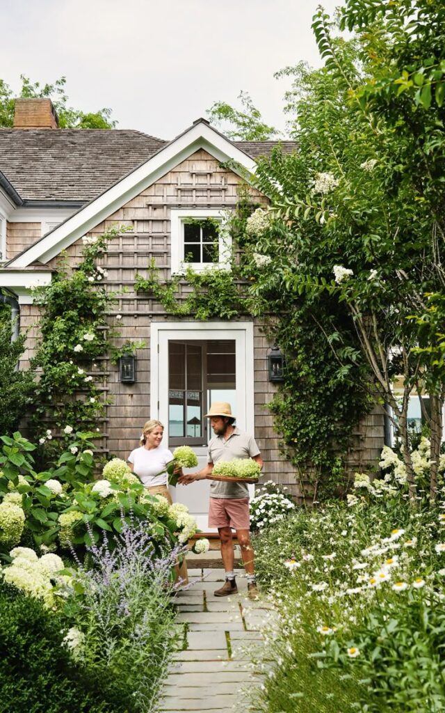 A couple gardening with hydrangeas in front of a charming cottage surrounded by lush greenery and blooming flowers.