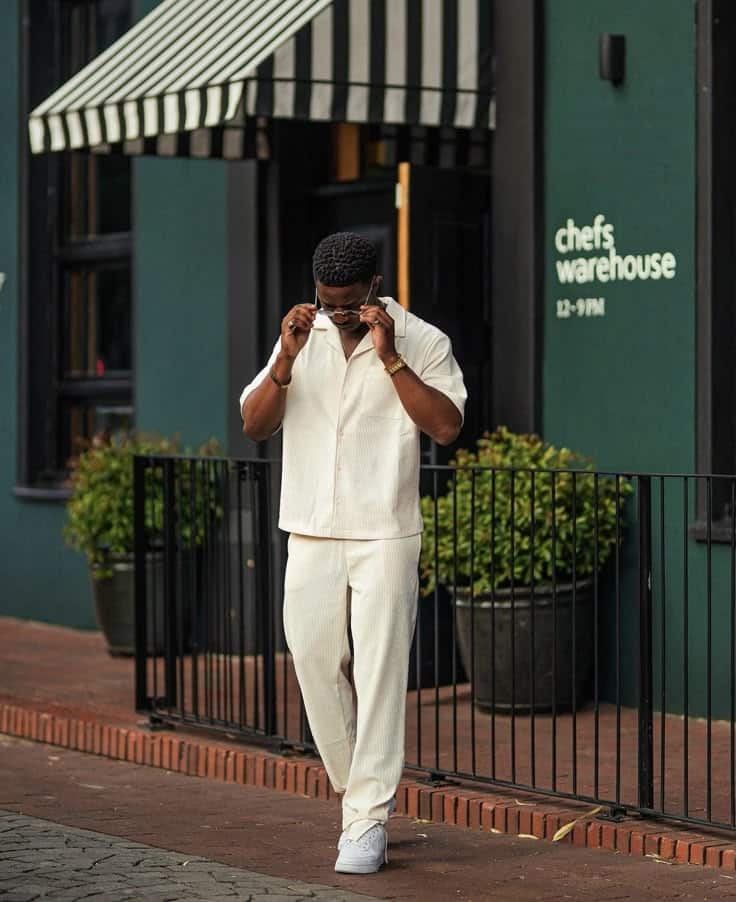 Man in a stylish cream outfit adjusts sunglasses while walking outside a restaurant with striped awnings.