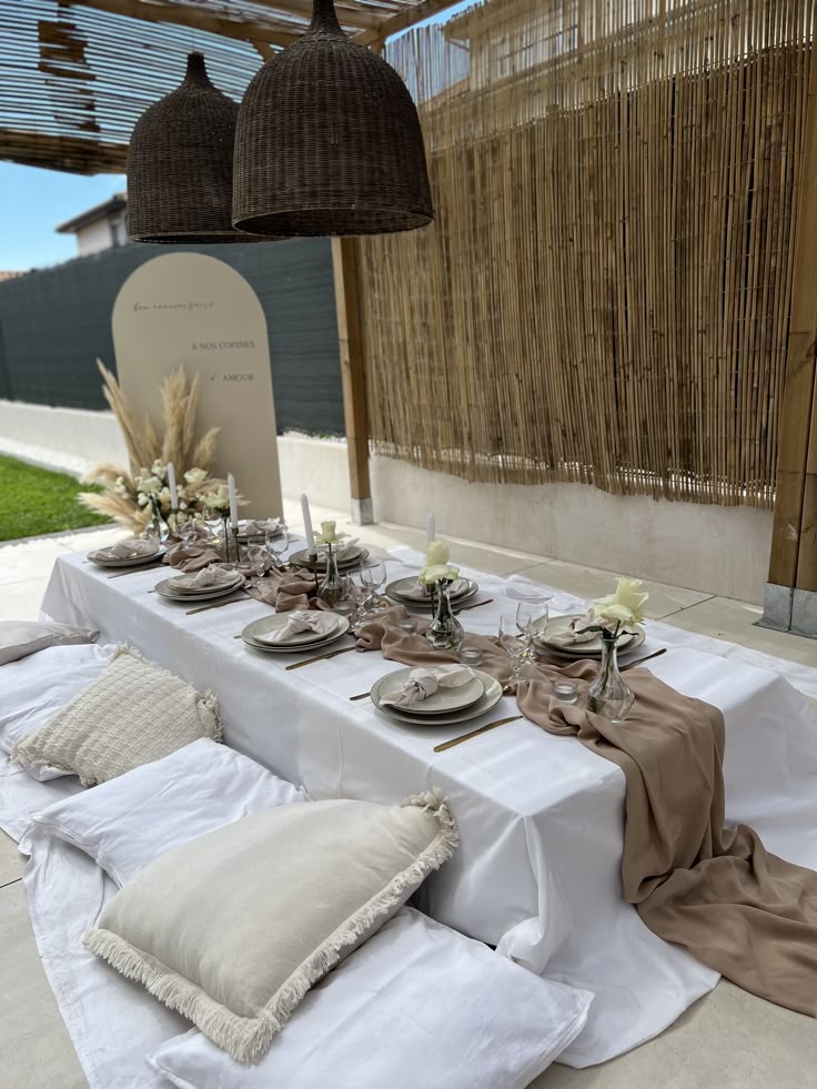 Outdoor elegant dining setup with white tablecloth, beige decor, candles, and pillows under wicker lampshades.