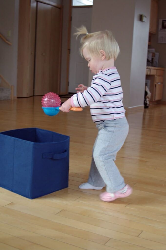 Toddler playing with a toy next to a blue storage bin on a wooden floor indoors.