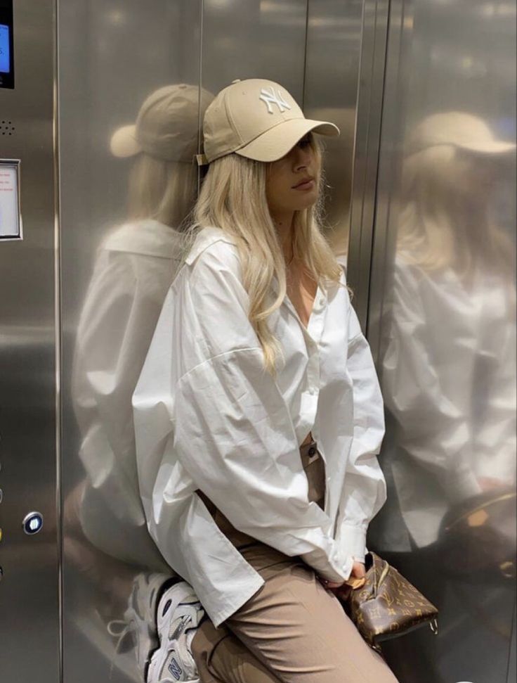 Blonde woman in a white shirt and baseball cap, leaning against an elevator wall with her reflection visible.
