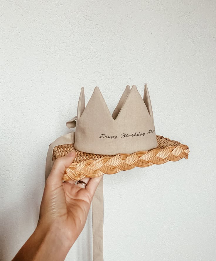Hand holding a fabric birthday crown on a woven stand against a white wall.