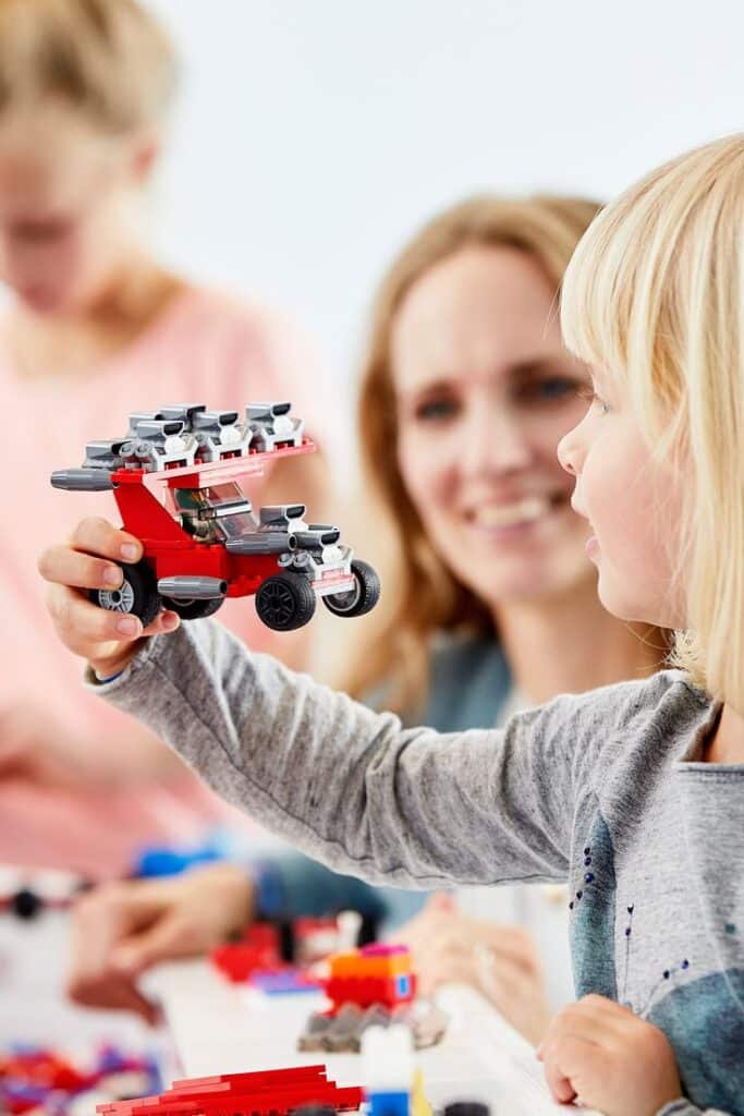 Blue Zone - Test Driver race car Child holding a LEGO car, with a smiling woman in the background. | Sky Rye Design Child holding a LEGO car, with a smiling woman in the background.