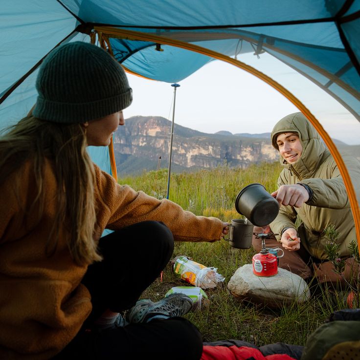 Two campers inside a tent cooking on a portable stove with scenic mountain views in the background.