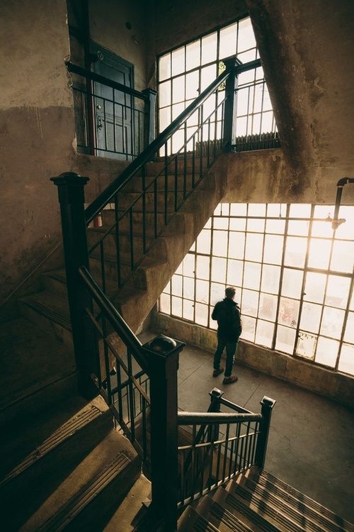 Person standing by large industrial windows in vintage stairway, gazing outside at sunset. Industrial building interior.