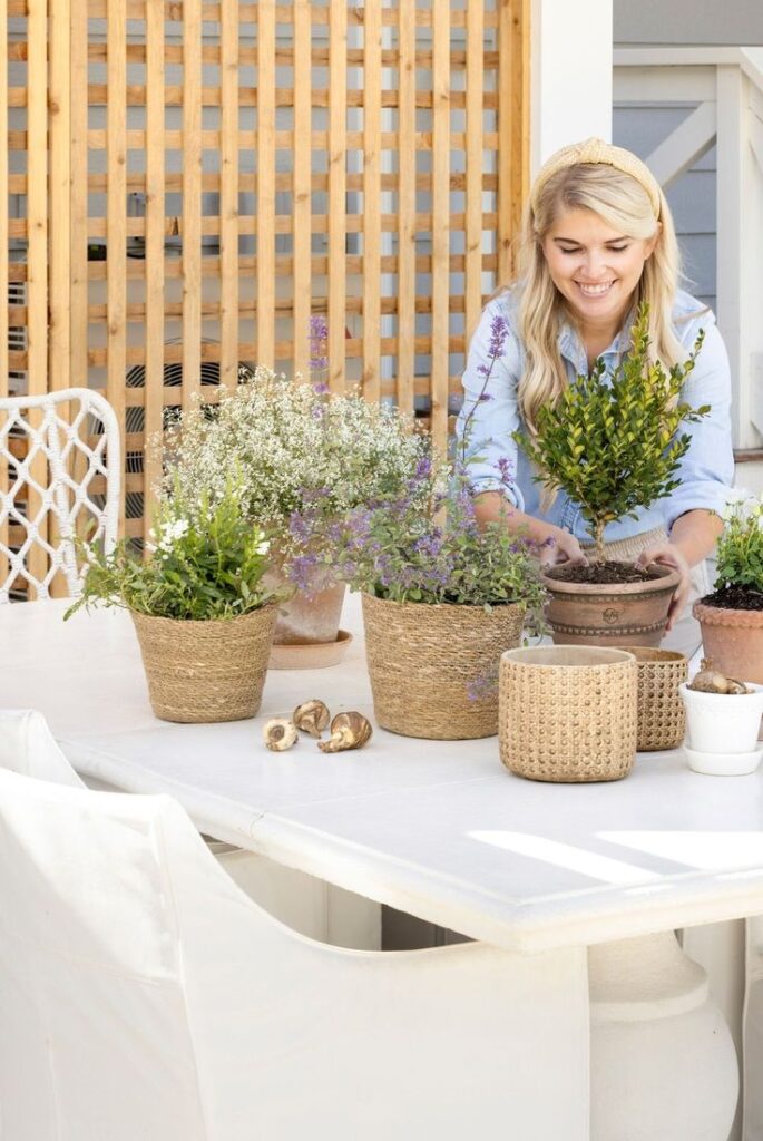 Person arranging indoor plants on table, surrounded by woven pots, smiling and enjoying gardening.