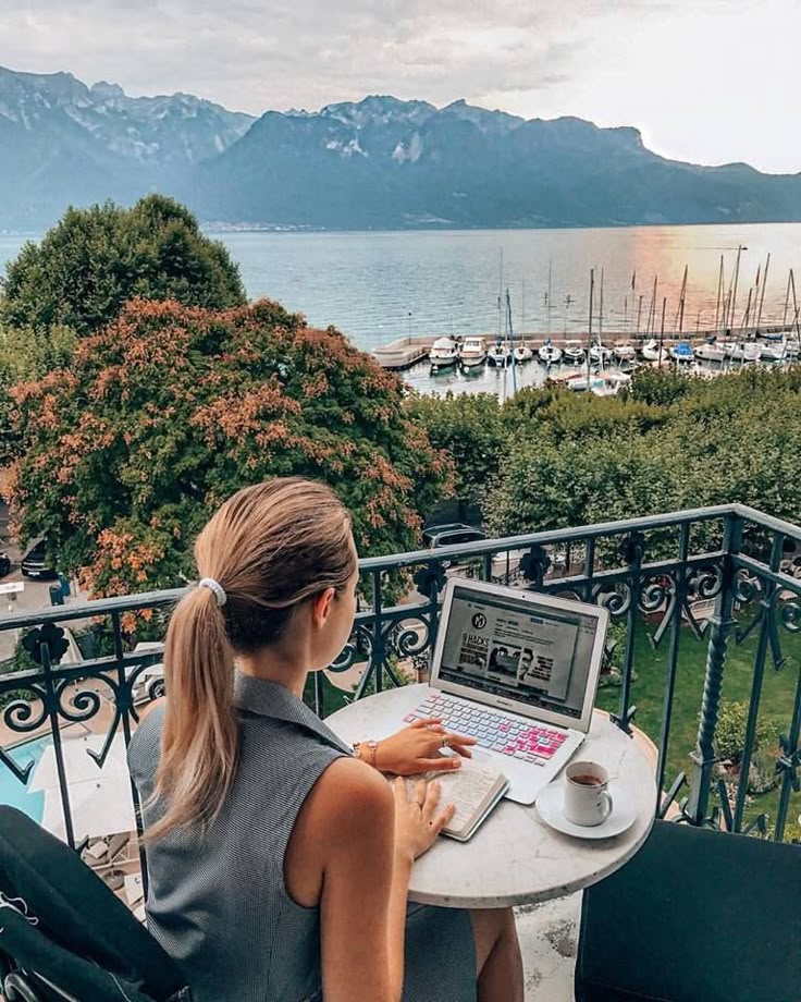 Person working on a laptop with a scenic mountain and lake view from a balcony, enjoying coffee.