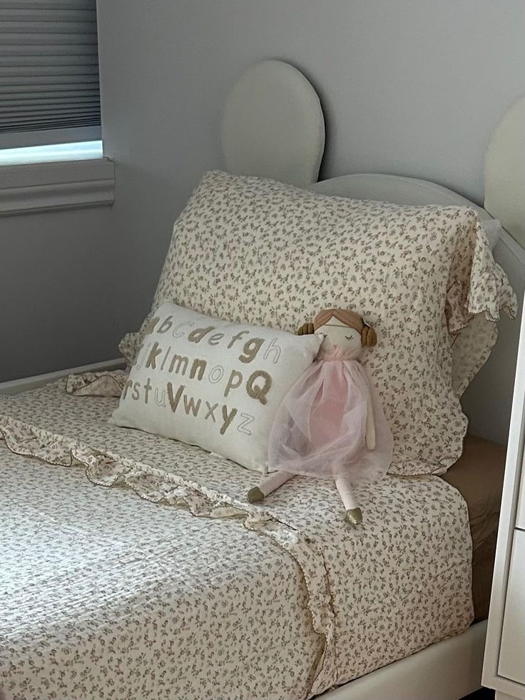 Children's bed with floral bedding, alphabet pillow, and a doll in a pink dress. Cozy and decorative setup.