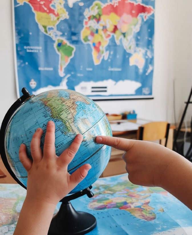Child pointing at a globe with a world map in the background, exploring geography in a classroom setting.